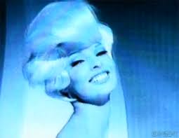 A blue-toned close-up portrait of a smiling woman with short light hair against a dark blue background.