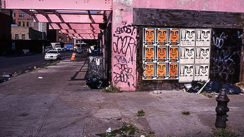 Urban street scene with a pink brick building covered in graffiti, a boarded window with orange-and-white decorative panels, an underpass structure overhead, parked cars, and a fire hydrant in the foreground.
